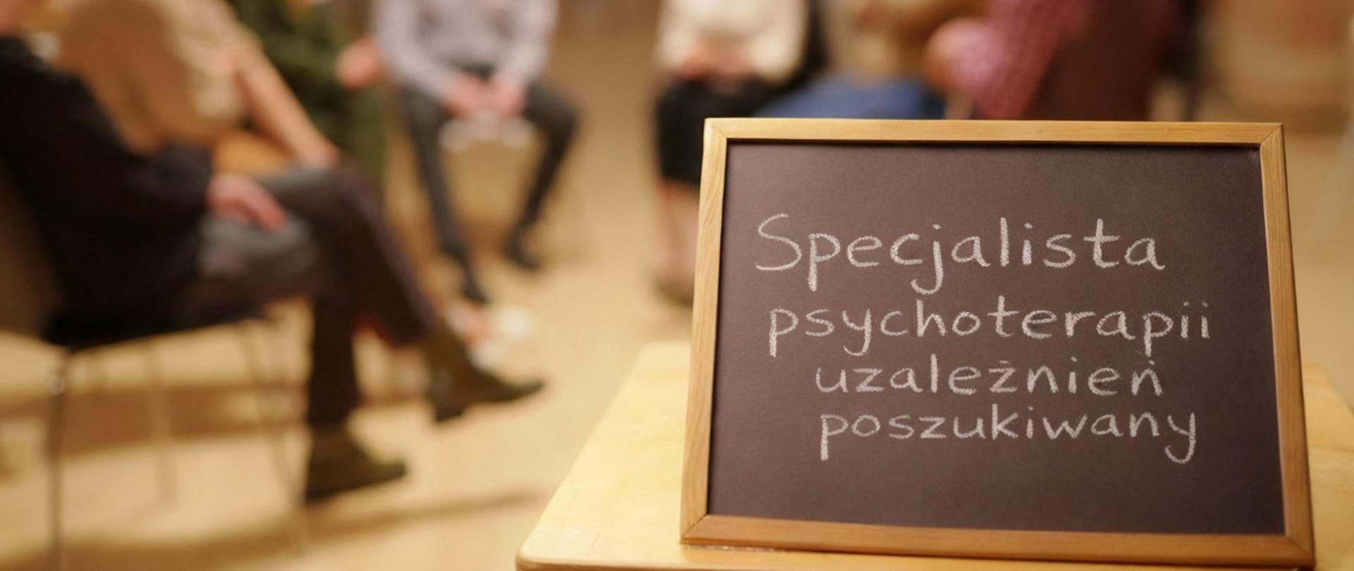 Tablica z napisem ‘Specjalista psychoterapii uzależnień poszukiwany’ na pierwszym planie. W tle grupa osób siedzących w kręgu podczas spotkania terapeutycznego.