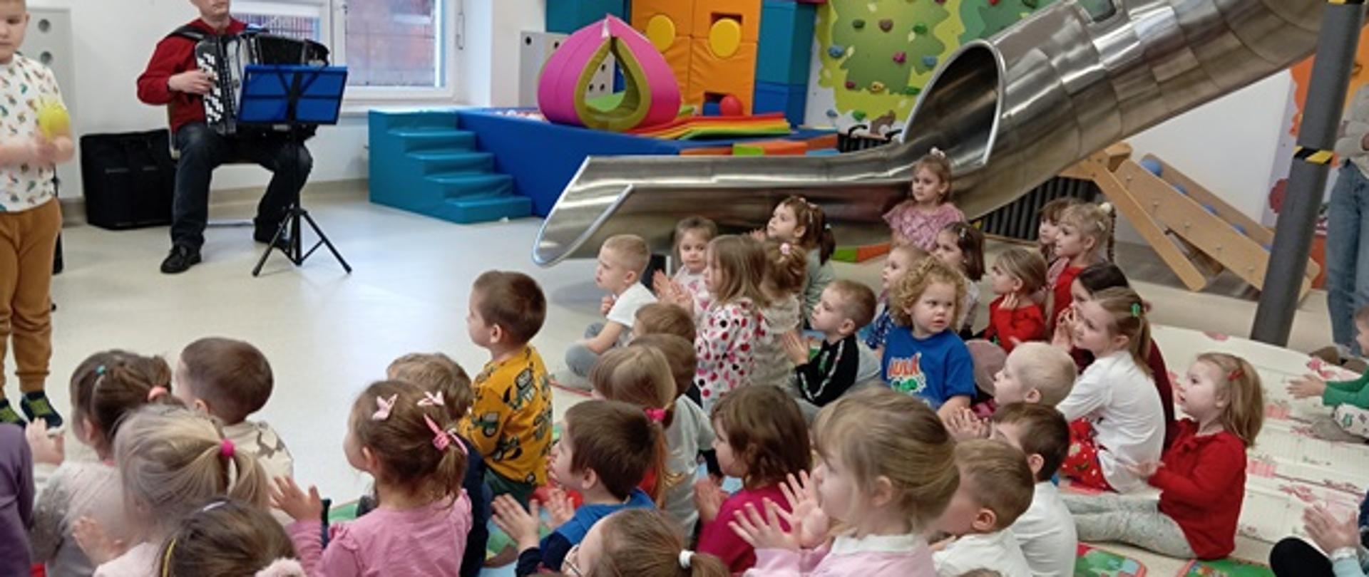 Zdjęcie prezentuje dzieci biorące udział w audycji muzycznej oraz teatrzyku. Przed grupą siedzących dzieci występują aktorzy