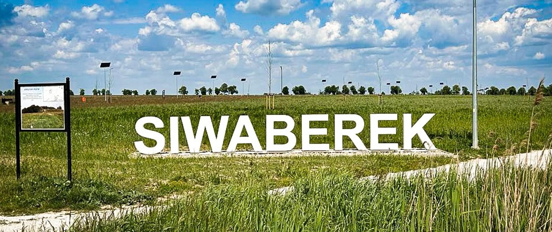 Panorama zielonego terenu z napisem „SIWABEREK” ustawionym z dużych białych liter na tle nieba z białymi chmurami. Obok znajduje się tablica informacyjna i maszt z panelem solarnym oraz turbiną wiatrową. W oddali widać kolejne słupy z panelami oraz drzewa na horyzoncie.