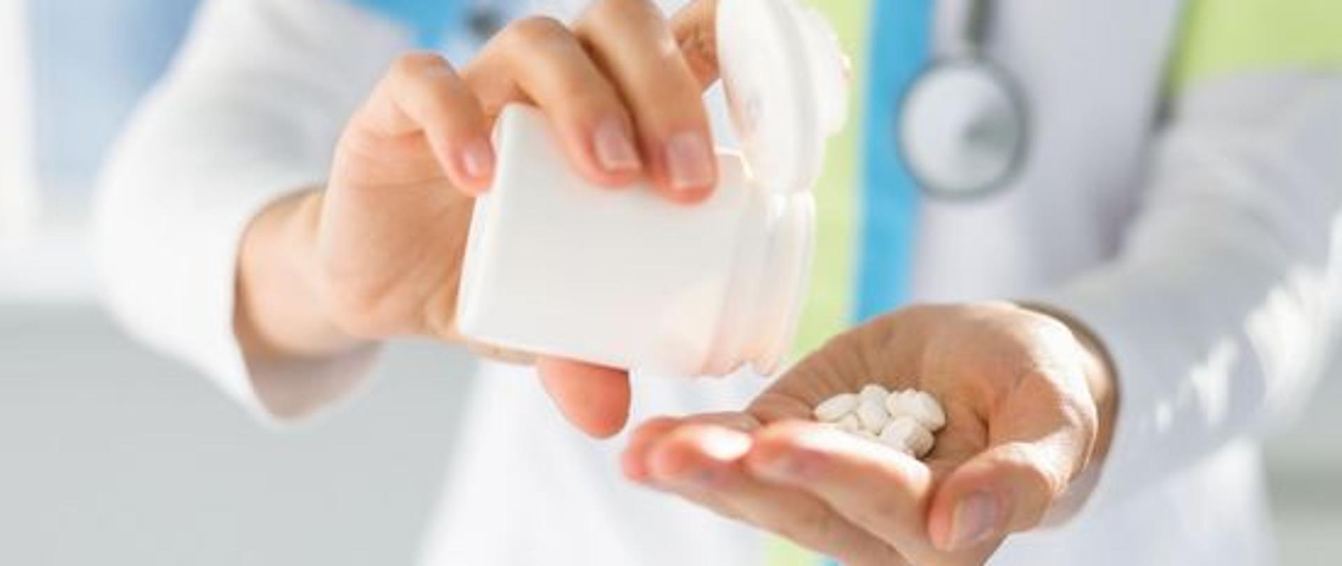 Na dłoni lekarza wysypane zostały białe tabletki. Pod zdjęciem napis "Amantadyna proces badań klinicznych związanych z leczeniem"