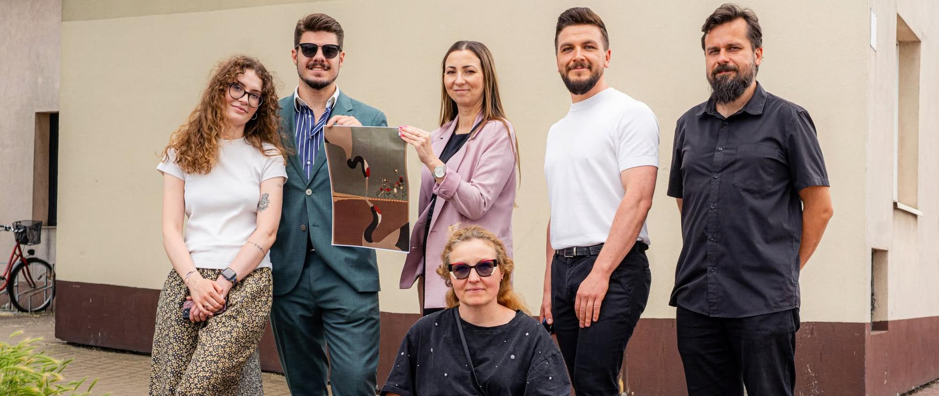 Jury pozuje do wspólnego zdjęcia przed budynkiem. Jedna z osób prezentuje nagrodzoną pracę przedstawiającą żurawie i motyw zszywania horyzontu.