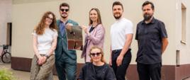 Jury pozuje do wspólnego zdjęcia przed budynkiem. Jedna z osób prezentuje nagrodzoną pracę przedstawiającą żurawie i motyw zszywania horyzontu.