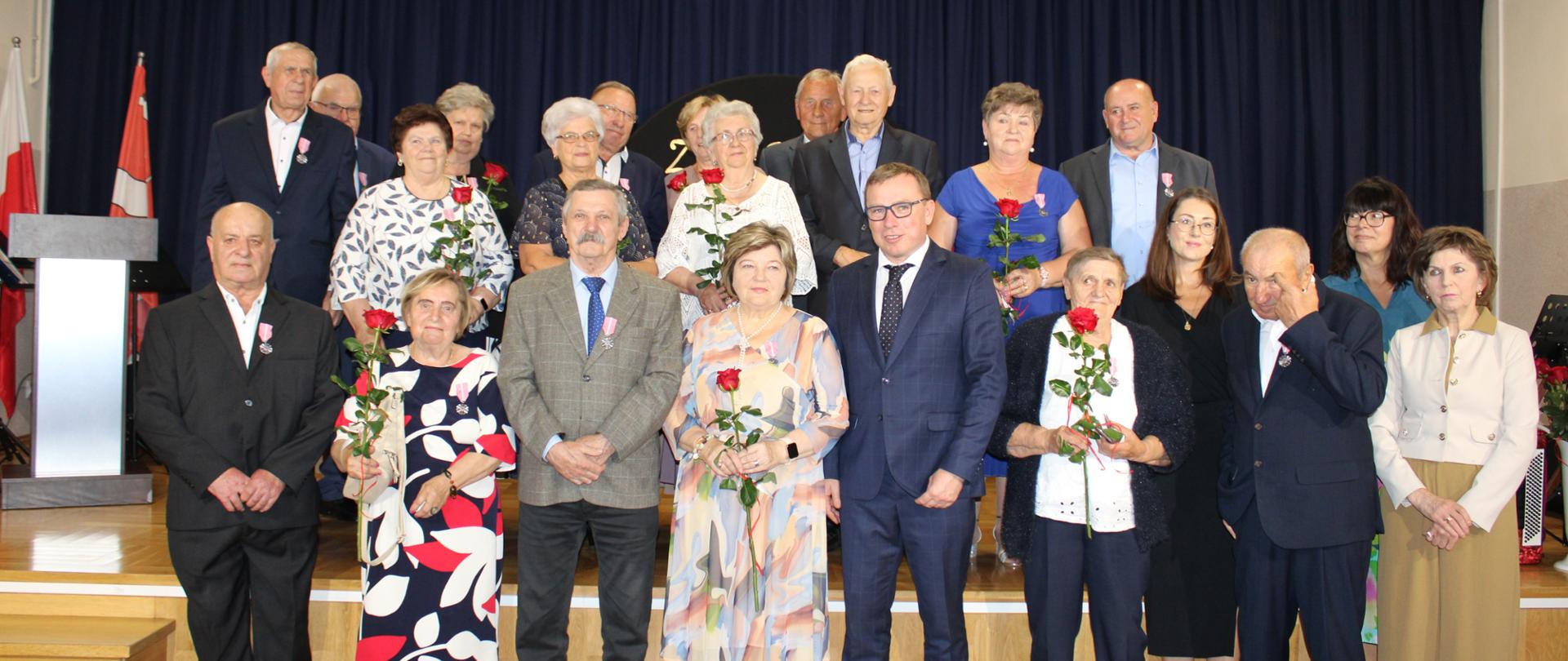 Grupowe zdjęcie par jubileuszowych z przedstawicielem urzędu na scenie udekorowanej napisem Złote Gody
