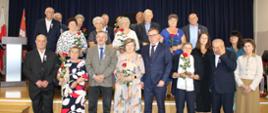 Grupowe zdjęcie par jubileuszowych z przedstawicielem urzędu na scenie udekorowanej napisem Złote Gody