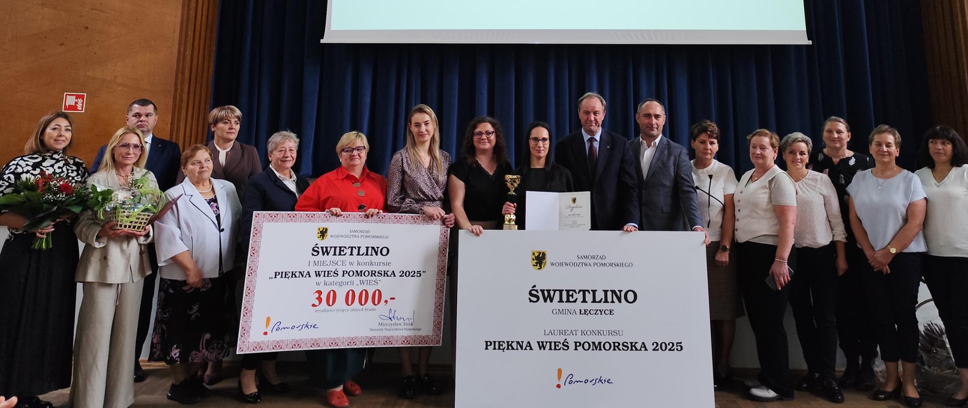 Osiemnaście osób pozujących do zdjęcia z tablicami informującymi o przyznaniu miejscowości Świetlino nagrody w wysokości 30 tysięcy złotych za zajęcie I miejsca w konkursie Piękna Wieś Pomorska 2025