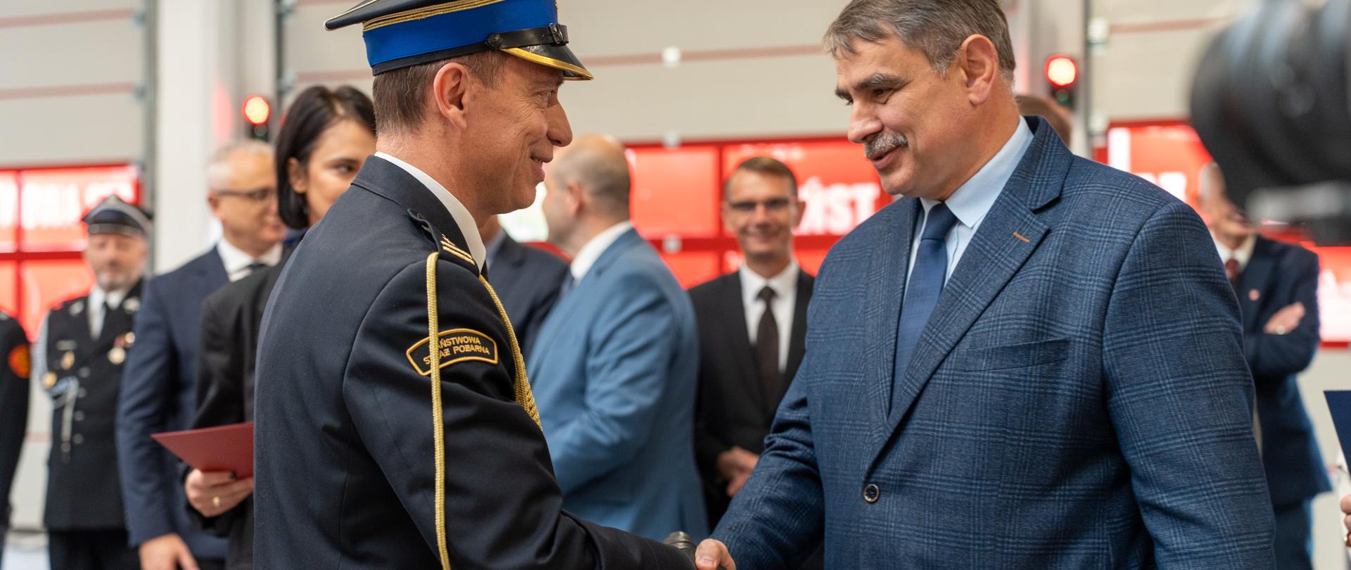 Burmistrz Miasta Kalwarii Zebrzydowskiej Tadeusz Stela składa gratulacje i ściska dłoń st. bryg. Wojciechowi Grzybczykowi podczas uroczystości przyjęcia obowiązków Komendanta Powiatowego Państwowej Straży Pożarnej w Wadowicach, która odbyła się 13 października 2026 roku. W tle widoczni są uczestnicy uroczystości, w tym przedstawiciele samorządów i służb mundurowych.