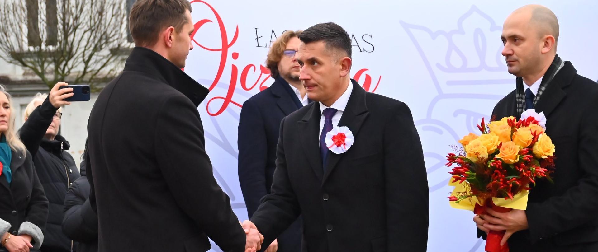 Dwóch mężczyzn w czarnych płaszczach podaje sobie dłonie podczas ceremonii. Jeden z nich trzyma bukiet kwiatów. W tle widoczny baner z napisem „Łączy nas Ojczyzna”.