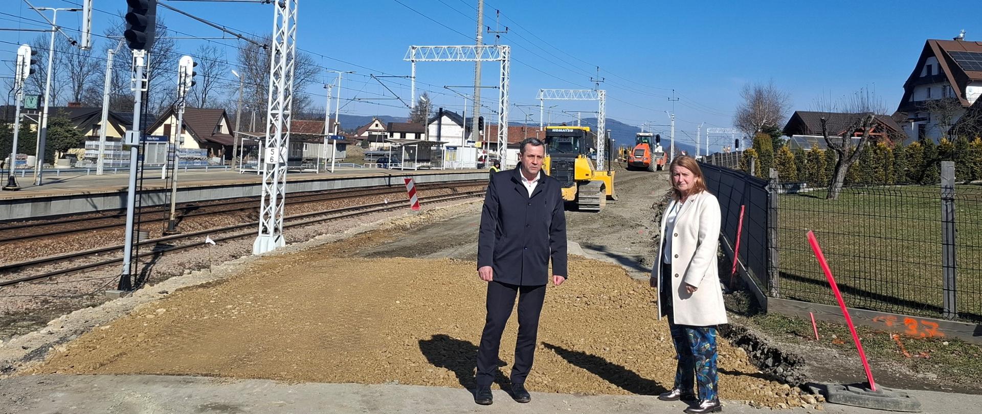 Dwie dorosłe osoby na placu budowy drogi, W tle tory kolejowe oraz maszyny budowlane