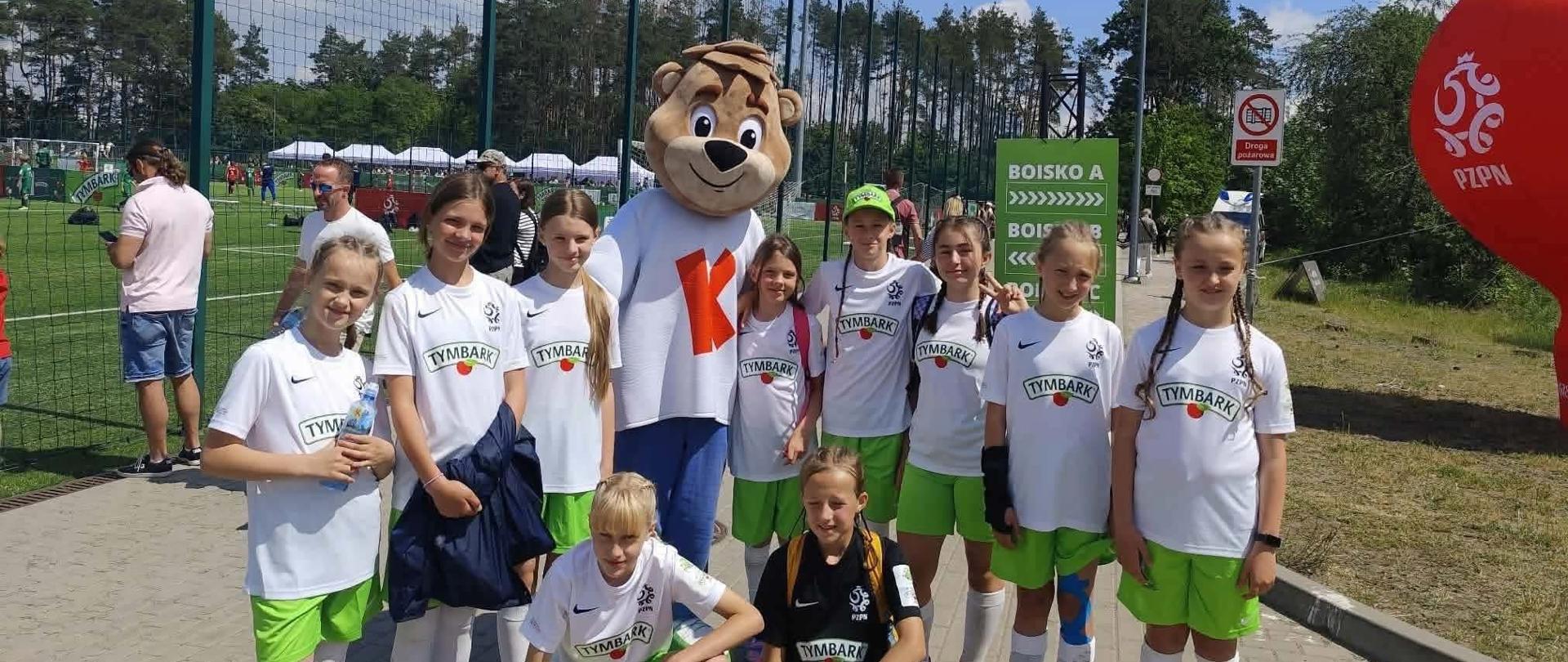 Drużyna dzieci w białych koszulkach pozuje do wspólnego zdjęcia na świeżym powietrzu podczas finału Ogólnopolskiego Pucharu Tymbarku. W tle znajduje się ogrodzenie boiska i balon reklamowy