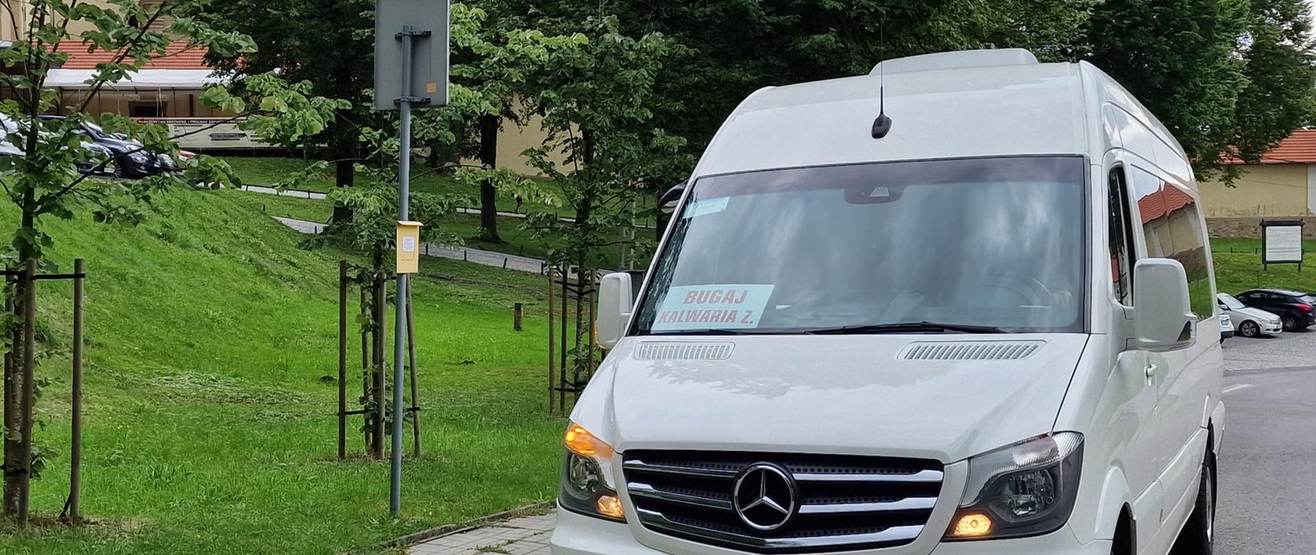 Biały bus marki Mercedes stoi z włączonym prawym kierunkowskazem przy chodniku na przystanku. Za nim drzewa, trawa, zabudowania klasztorne.