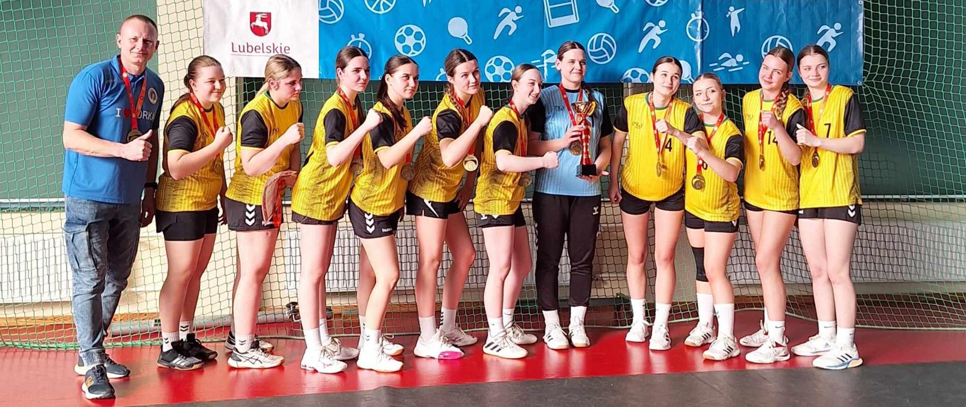 Dziewczęta z Zespołu Szkół nr 1 w Kraśniku stoją na hali sportowej w strojach sportowych z zawieszonymi na szyjach medalami. Pierwszy od lewej stoi nauczyciel wychowania fizycznego Mariusz Sielski. Nad dziewczętami zawieszony jest baner Szkolnego Związku Sportowego z napisem: Wojewódzkie Współzawodnictwo Szkół, Finał Wojewódzki w piłce ręcznej dziewcząt, Kraśnik 2025