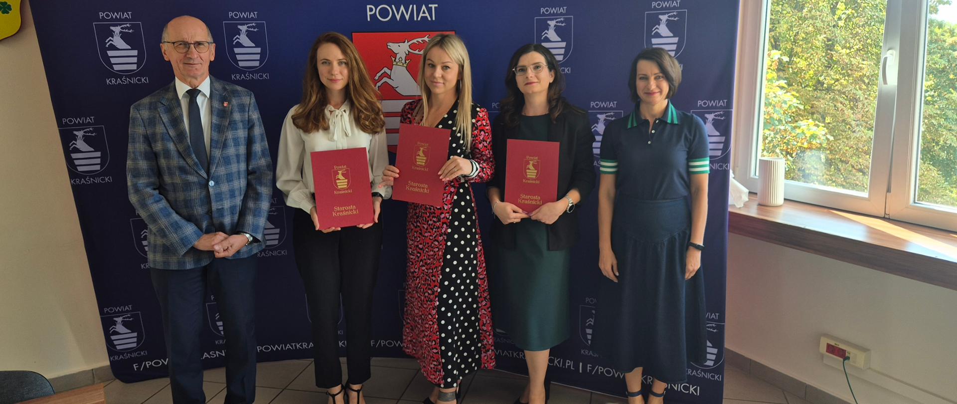 Na tle granatowej ścianki reklamowej Powiatu Kraśnickiego stoją trzy nauczycielki trzymające przed sobą w rękach bordowe teczki z aktami nadania stopnia awansu zawodowego nauczyciela mianowanego. Od ich lewej strony stoi Starosta Kraśnicki Zbigniew Dżugaj, zaś od prawej Kierownik Wydziału Edukacji i Sportu Starostwa Powiatowego w Kraśniku Agnieszka Drozd.