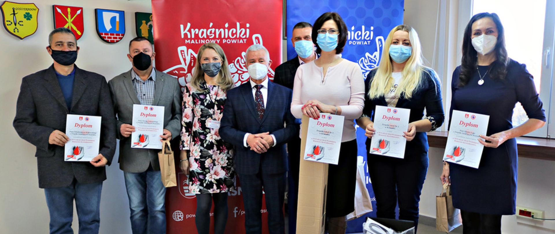 Na zdjęciu znajduje się osiem osób, cztery kobiety i czterech mężczyzn. Po środku stoją Starosta Kraśnicki Andrzej Rolla i kierownik wydziału Spraw Społecznych. Dyrektorzy szkół oraz opiekunowie trzymają podziękowania w postaci dyplomów, a także nagrody dla swoich uczniów, którzy włączyli się w akcję honorowego krwiodawstwa. Dyplomy oraz nagrody wręczył Starosta Kraśnicki Andrzej Rolla.