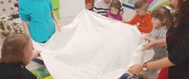 Dzieci w trakcie świątecznych zabaw - wspólnie trzymają białą tkaninę nad leżącą osobą
