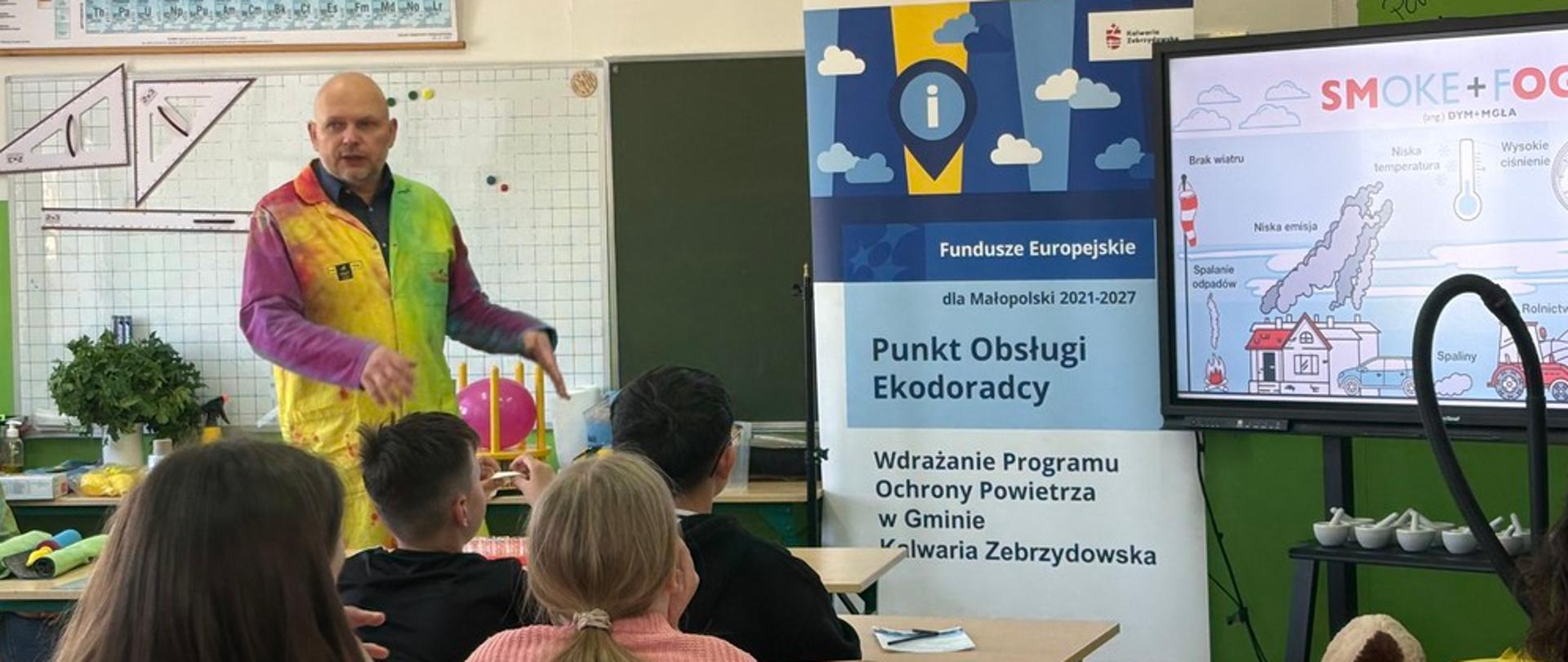 Szkolna sala. Uczniowie siedzący w ławkach oglądają prezentację na temat smogu, prowadzoną przez mężczyznę ubranego w kolorowy strój.