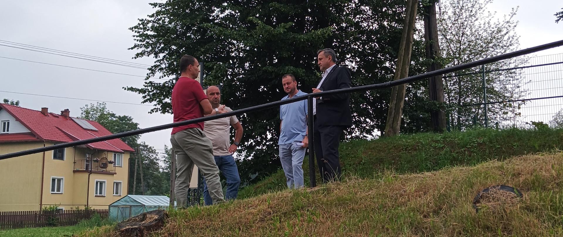 Czterech mężczyzn stoi na trawiastym wzniesieniu i rozmawia przy metalowej barierce. W tle rośnie duże, gęste drzewo, obok znajduje się słup energetyczny oraz ogrodzenie. Po lewej stronie widoczne są domy jednorodzinne z czerwonymi dachami i niewielka szklarnia. Niebo jest pochmurne, a teren sprawia wrażenie wiejskiej lub podmiejskiej okolicy.