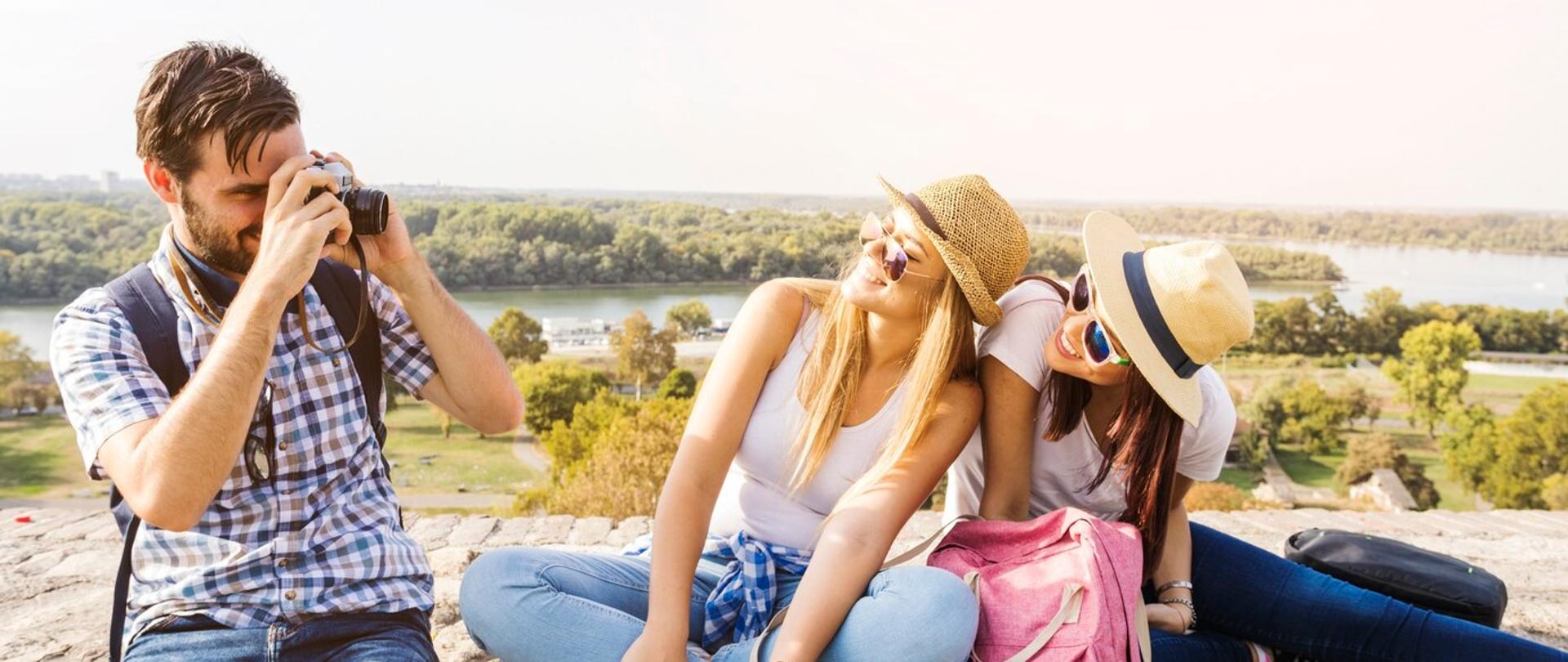 Mężczyzna fotografuje dwie kobiety podczas wspólnych wakacji, siedzą na kamiennym murku, w tle widać lasy i jezioro. Ubrani są typowo wakacyjnie, kobiety mają mapkę rozłożoną