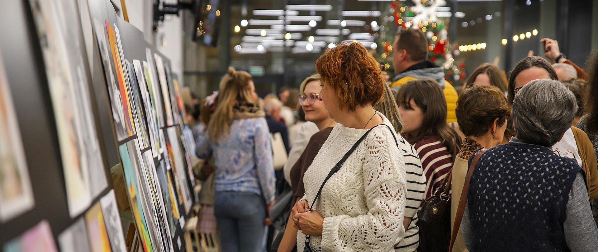 W jasnej, przestronnej sali odbywa się wernisaż. Po lewej stronie ustawiono rzędy sztalug z kolorowymi pracami plastycznymi. Wzdłuż nich przechadza się wiele osób, oglądając wystawione prace. Na pierwszym planie stoi kobieta w białym, ażurowym swetrze, trzymająca czarną torebkę na ramieniu i uważnie przyglądająca się obrazom. W tle widać tłum gości oraz ozdobioną choinkę, tworzącą świąteczną atmosferę. 