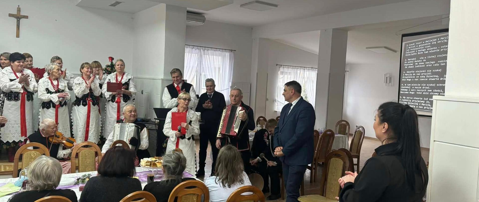 Wnętrze sali podczas spotkania opłatkowego. Przy stołach siedzą uczestnicy wydarzenia, a przed nimi występuje zespół ludowy w tradycyjnych strojach, śpiewający kolędy i grający na instrumentach, w tym na akordeonie i skrzypcach. Obok zespołu stoją zaproszeni goście, w tym burmistrz w garniturze, słuchający występu. Na ścianie widoczny krzyż, a atmosfera spotkania jest uroczysta i serdeczna.
