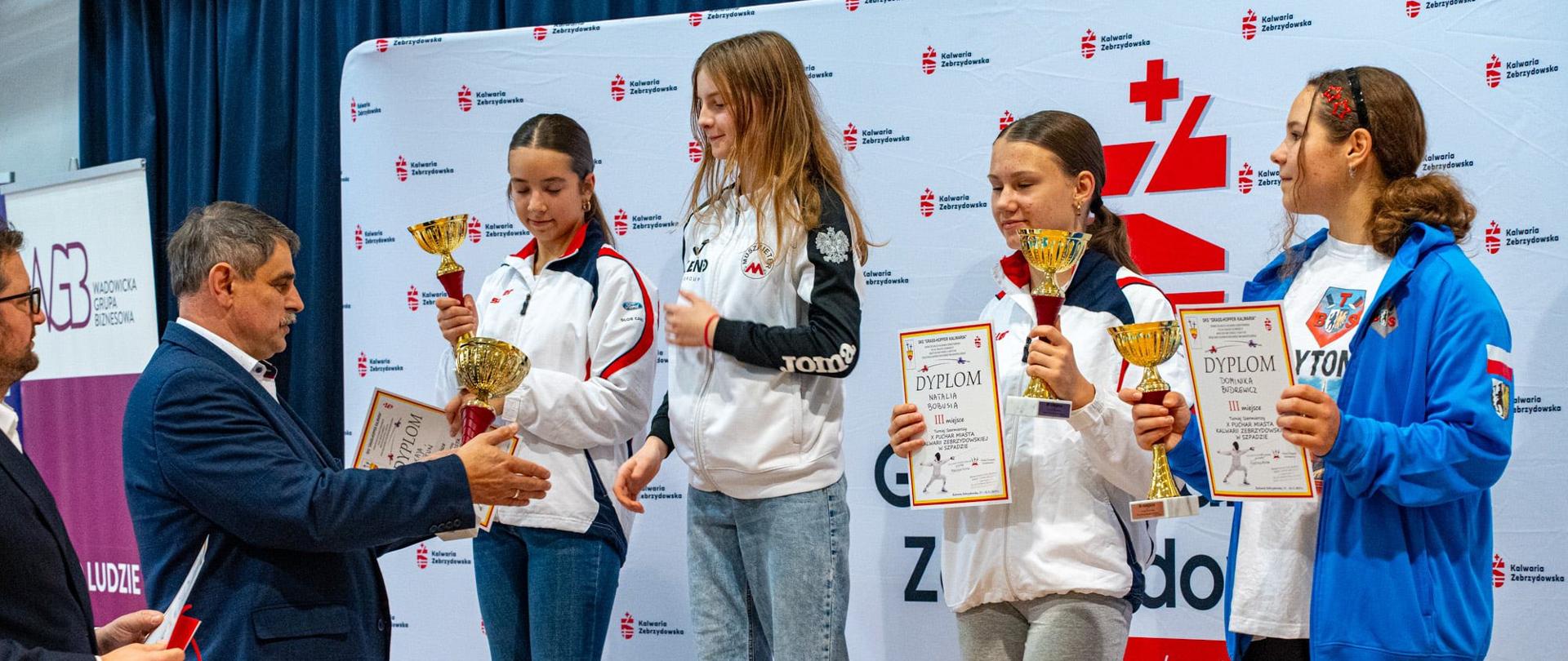 Na podium stoją cztery młode zawodniczki w sportowych strojach, trzymając puchary i dyplomy za zajęte miejsca w zawodach. Przed podium Burmistrz Kalwarii Zebrzydowskiej Tadeusz Stela wręcza jednej z dziewcząt dyplom i nagrodę. Obok niego stoi mężczyzna z czerwoną torbą upominkową. W tle znajduje się ścianka z logo Gminy Kalwaria Zebrzydowska oraz grafika związana z wydarzeniem sportowym. Zawodniczki uśmiechają się i prezentują swoje trofea.