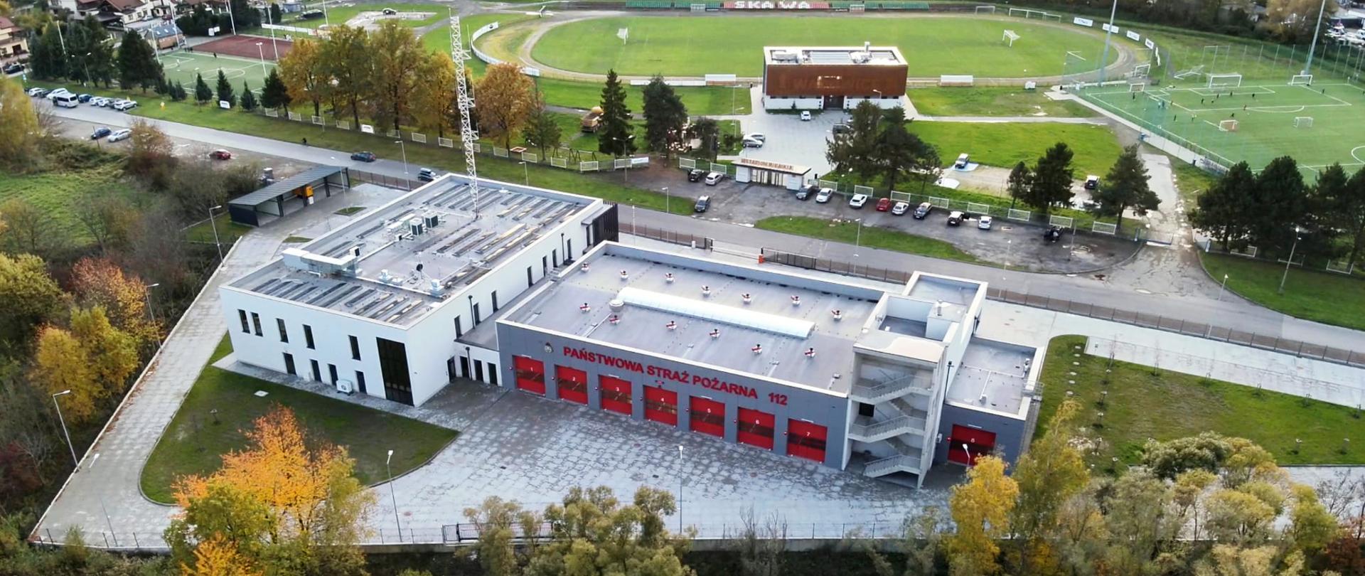 Zdjęcie przedstawia nowoczesny budynek Komendy Powiatowej Państwowej Straży Pożarnej w Wadowicach, sfotografowany z lotu ptaka. Obiekt wyróżnia się jasną elewacją, płaskim dachem z instalacjami technicznymi oraz czerwonymi bramami wjazdowymi do 14-stanowiskowego garażu. Przed budynkiem znajduje się szeroki plac manewrowy, a całość otoczona jest zadbaną zielenią i alejkami. W tle widoczne są kompleksy sportowe – stadion lekkoatletyczny, boiska piłkarskie oraz trybuny, a także zabudowa mieszkalna. Infrastruktura strażacka została zlokalizowana w strategicznym, dobrze skomunikowanym miejscu.