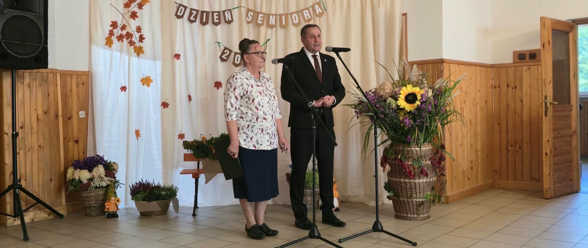 Dwie osoby na tle jesiennej ścianki z okazji dnia seniora, przemawiają przez mikrofon.