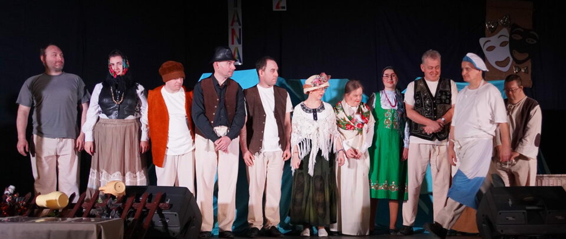 Grupa osób stoi na scenie w kostiumach podczas występu teatralnego.