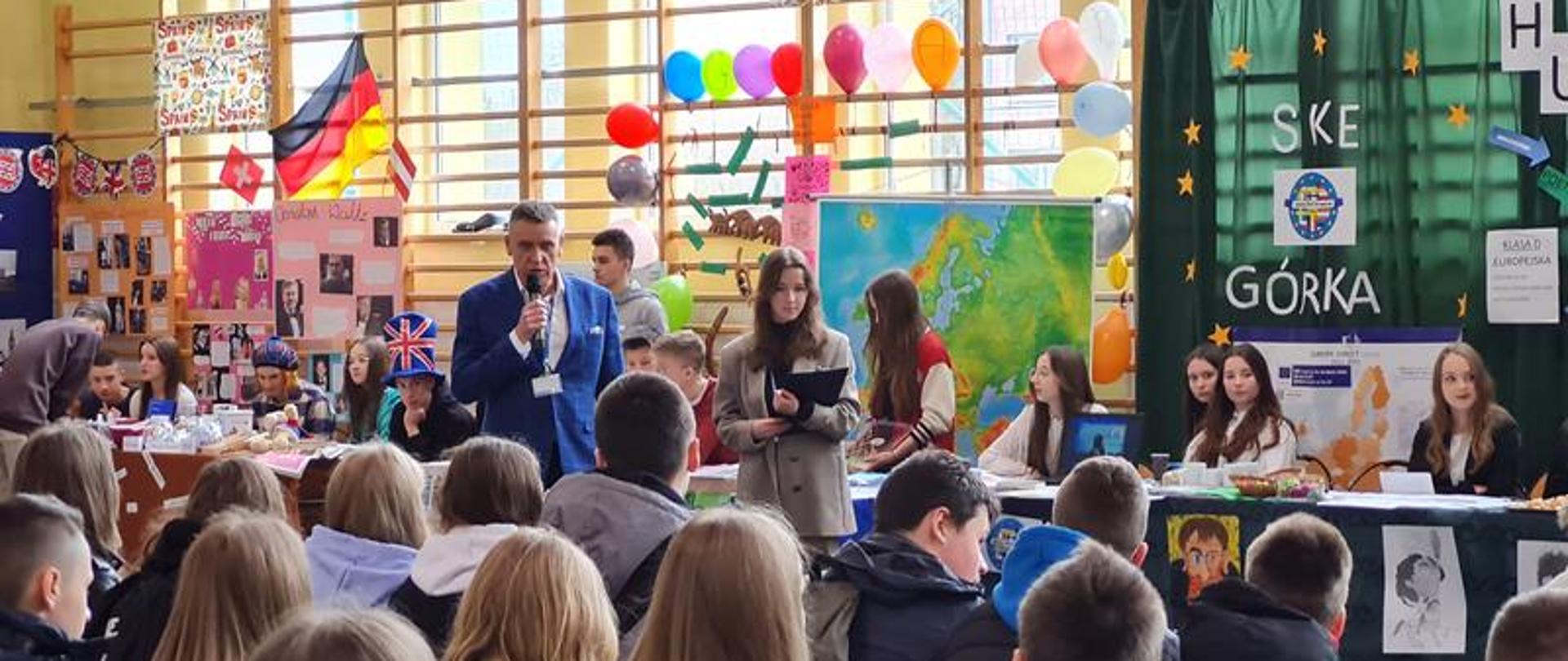 Zdjęcie przedstawia młodzież siedzącą na sali gimnastycznej Zespołu Szkół nr 1 w Kraśniku. Uczniowie szkoły siedzą przy stolikach stojących pod drabinkami, na których zawieszone są balony oraz plakaty promujące poszczególne szkoły. Naprzeciwko nich w ławkach siedzą uczniowie, którzy przybyli na Dni otwarte.