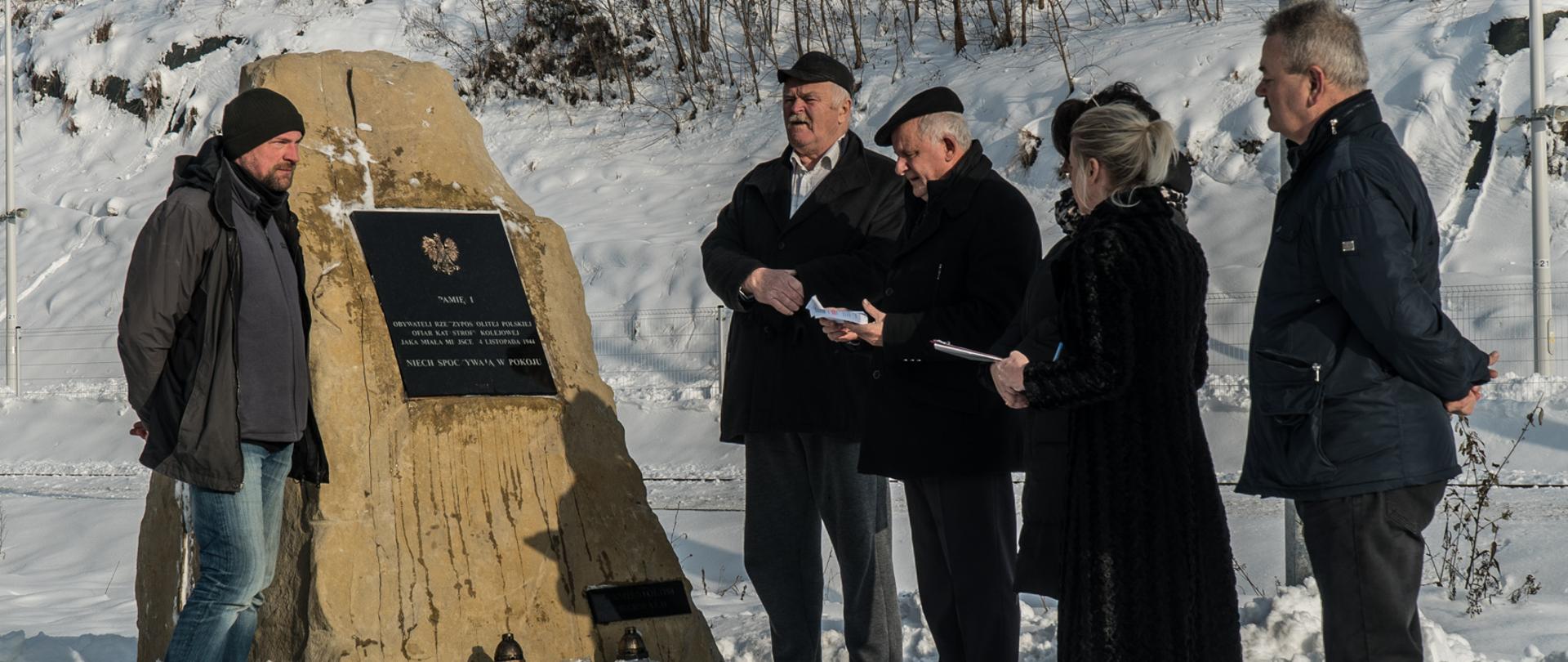 W zimowej aurze przy odnowionym obelisku stoi z lewej strony w kurtce i czapce mężczyzna z prawej strony widać trzech mężczyzn oraz kobietę w zimowym ubiorze.