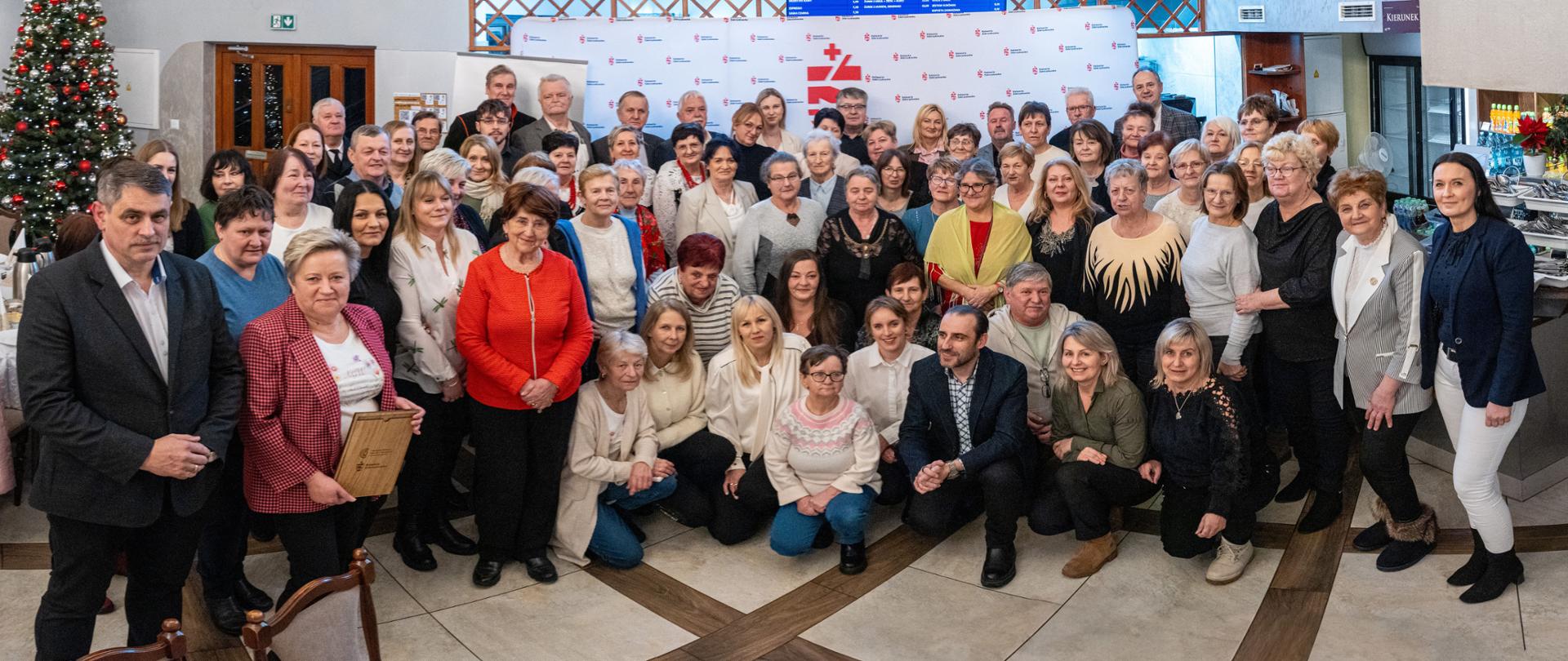 Zdjęcie grupowe kilkudziesięciu osób podczas spotkania w sali budynku publicznego. Uczestnicy stoją i klęczą w kilku rzędach, pozując do wspólnej fotografii. W tle widoczna jest ścianka z logotypami, udekorowana choinka oraz elementy wnętrza sali. Wśród uczestników znajdują się głównie kobiety, a spotkanie ma charakter oficjalno-integracyjny.