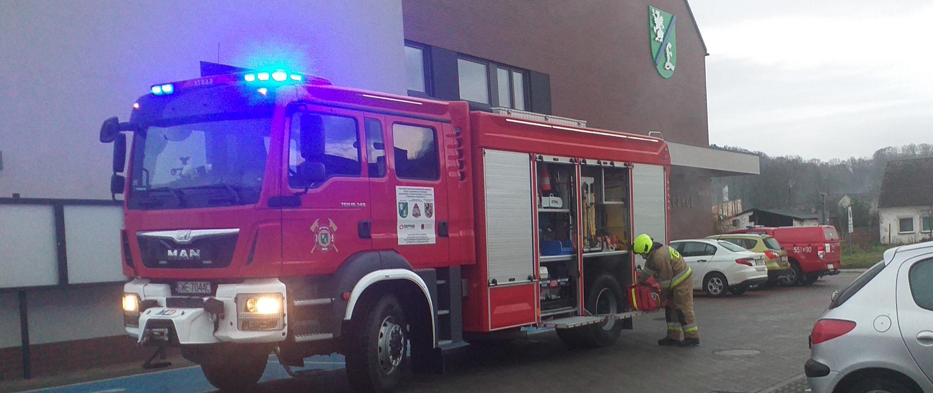 Zdjęcie strażaka wyciągającego torbę z wozu strażackiego przed budynkiem Urzędu Gminy