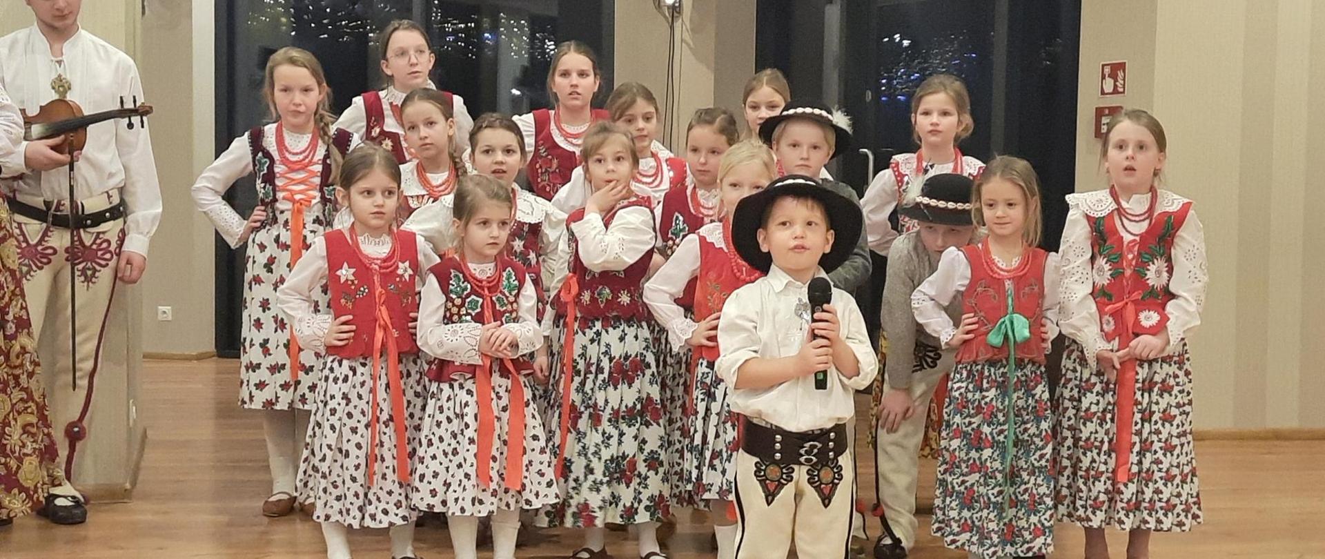 Dzieci z zespołu góralskiego, wszyscy w strojach góralskich w tle trzy rzędy dziewczynek z przodu solista z mikrofonem. 