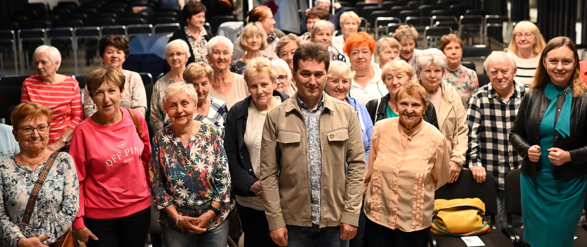 Zdjęcie grupowe uczestników spotkania seniorów
Grupa uśmiechniętych osób, głównie seniorek, pozuje do zdjęcia w jasnej sali z rzędami krzeseł w tle. Na pierwszym planie stoi mężczyzna w beżowej kurtce i kobieta w zielonej sukience z czarną kurtką. W tle widać inne osoby wychodzące z sali.