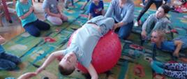 Ćwiczenia - chłopiec leży na plecach na piłce, nauczycielka go przytrzymuje, a dookoła siedzą dzieci
