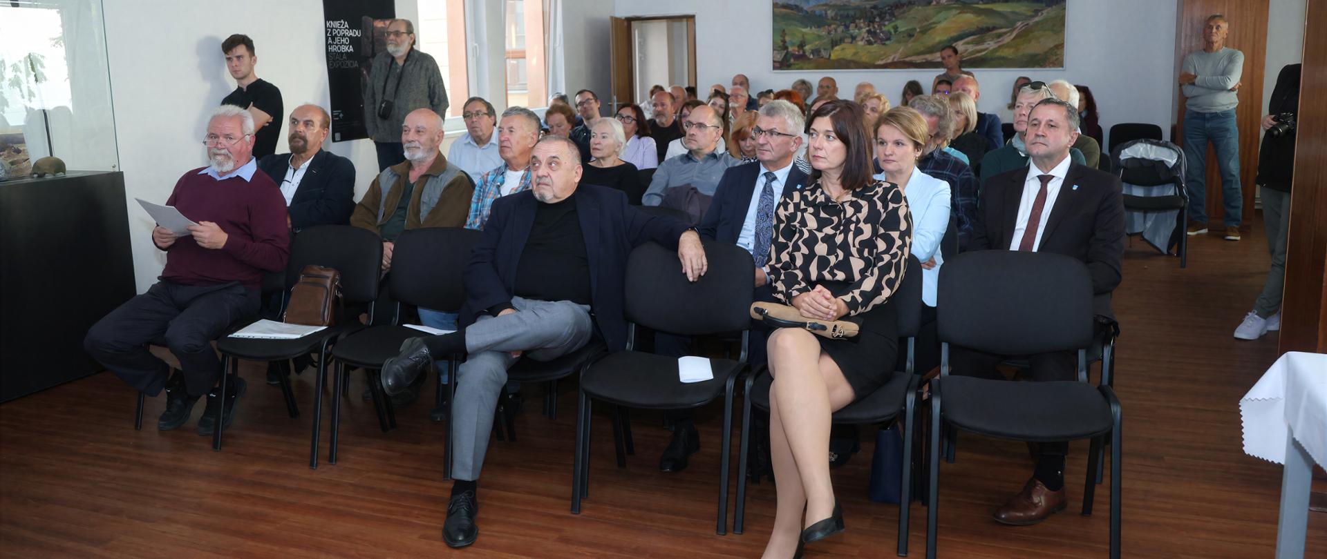 Grupa uczestników spotkania siedzi na krzesłach w sali konferencyjnej i pozuje do wspólnego zdjęcia.