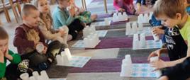 Dzieci w trakcie zabawy - siedzą w kółku, mają przed sobą kartki i kubki służące do zabawy