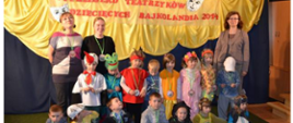 Grupa teatralna z trzema opiekunkami pozuje na tle dekoracji z napisem VI Przegląd Teatrzyków Dziecięcych Bajkolandia 2014