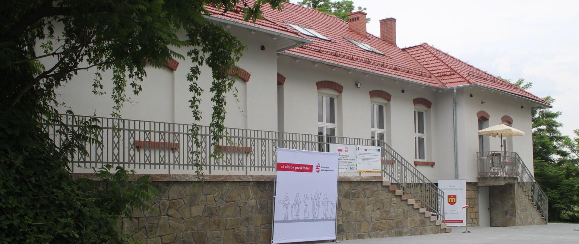 Budynek odrestaurowanego budynku, w lewej części drzewo, w centralnym budynek o jasnej elewacji, przykryty dachówką