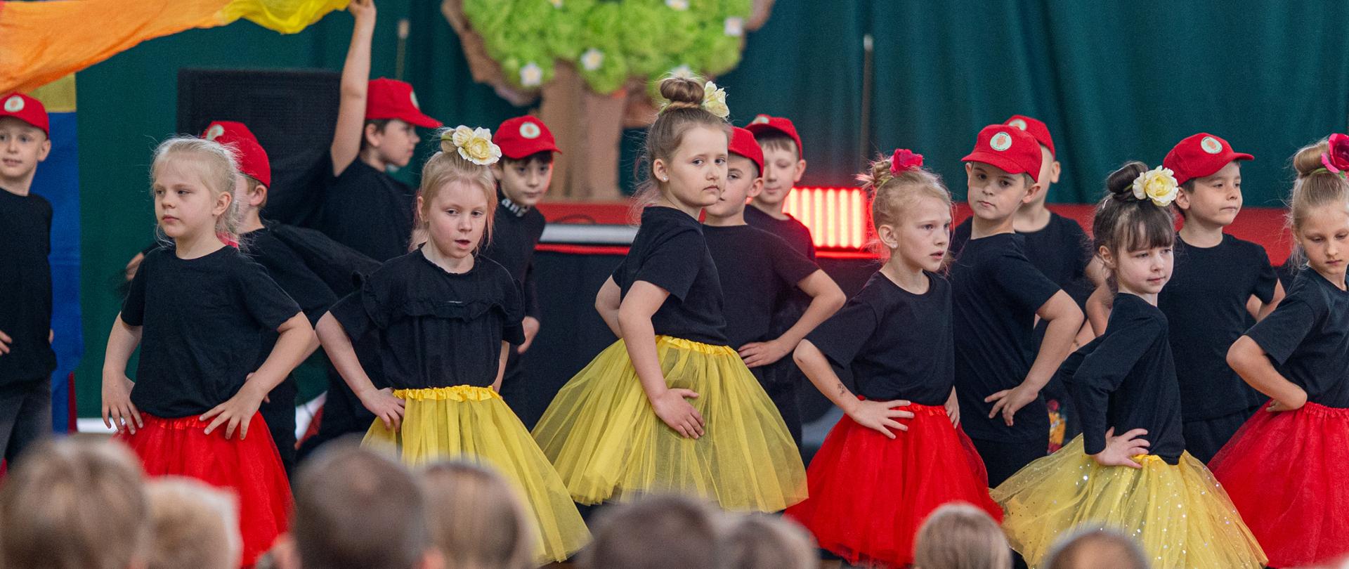 Dzieci w kolorowych strojach tanecznych występują na scenie podczas Festiwalu Kultur. Publiczność ogląda występ z widowni.
