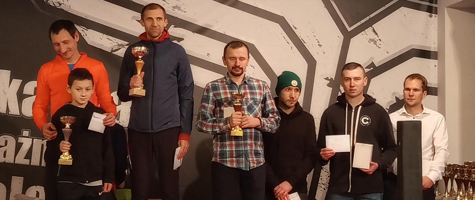 Zawodnicy stojący na podium wraz z pucharami i dyplomami. Na trzecim stopniu podium stoi Kamil Młynarz.