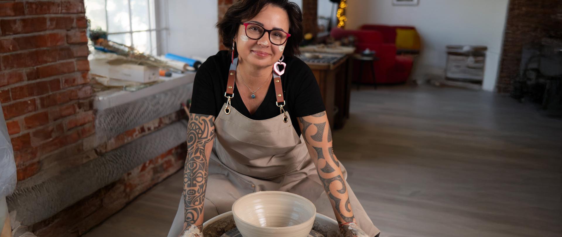 Kobieta siedzi przy kole garncarskim obok świeżo uformowanego ceramicznego naczynia.