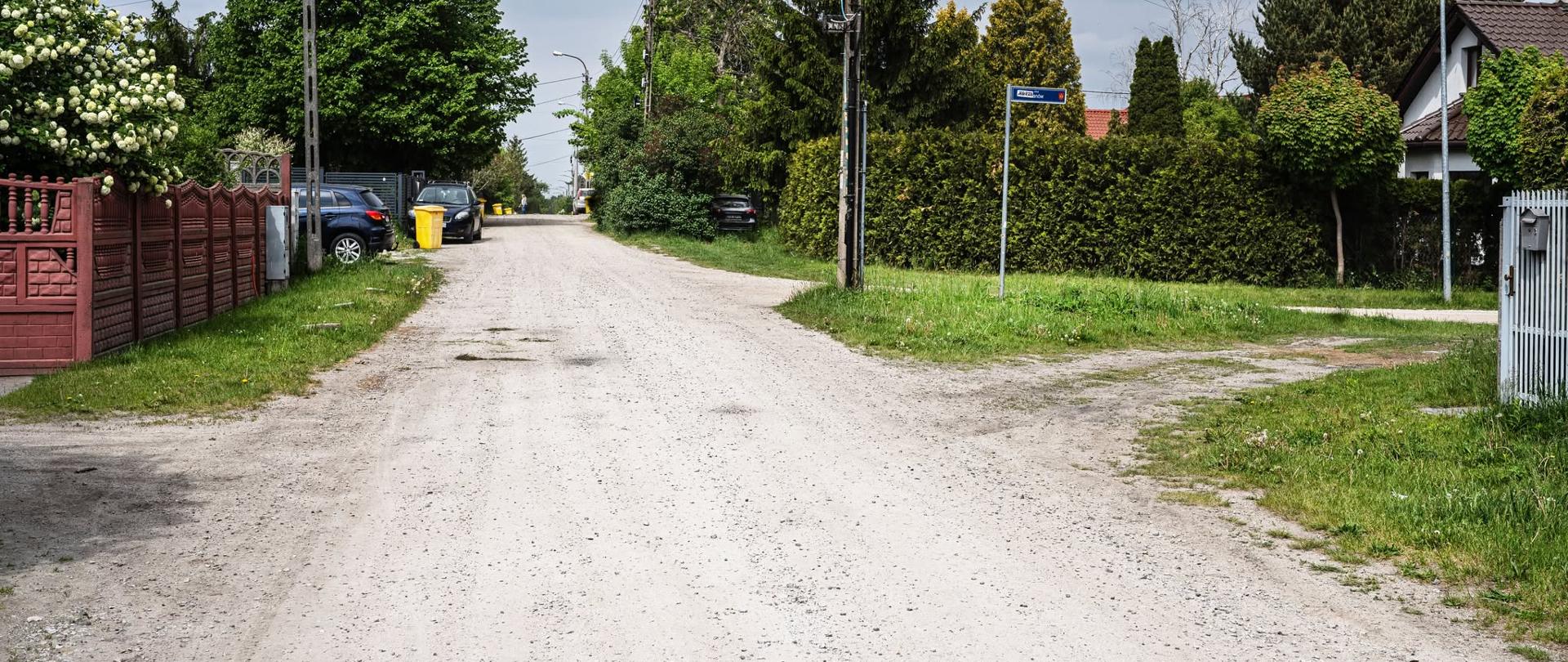 Nieutwardzona droga osiedlowa biegnąca między posesjami jednorodzinnymi. Po obu stronach znajdują się ogrodzenia, drzewa, krzewy oraz zaparkowane samochody. Wzdłuż ulicy stoją słupy energetyczne i latarnie, a przy skrzyżowaniu widoczna jest tablica z nazwą ulicy. Niebo jest częściowo zachmurzone.