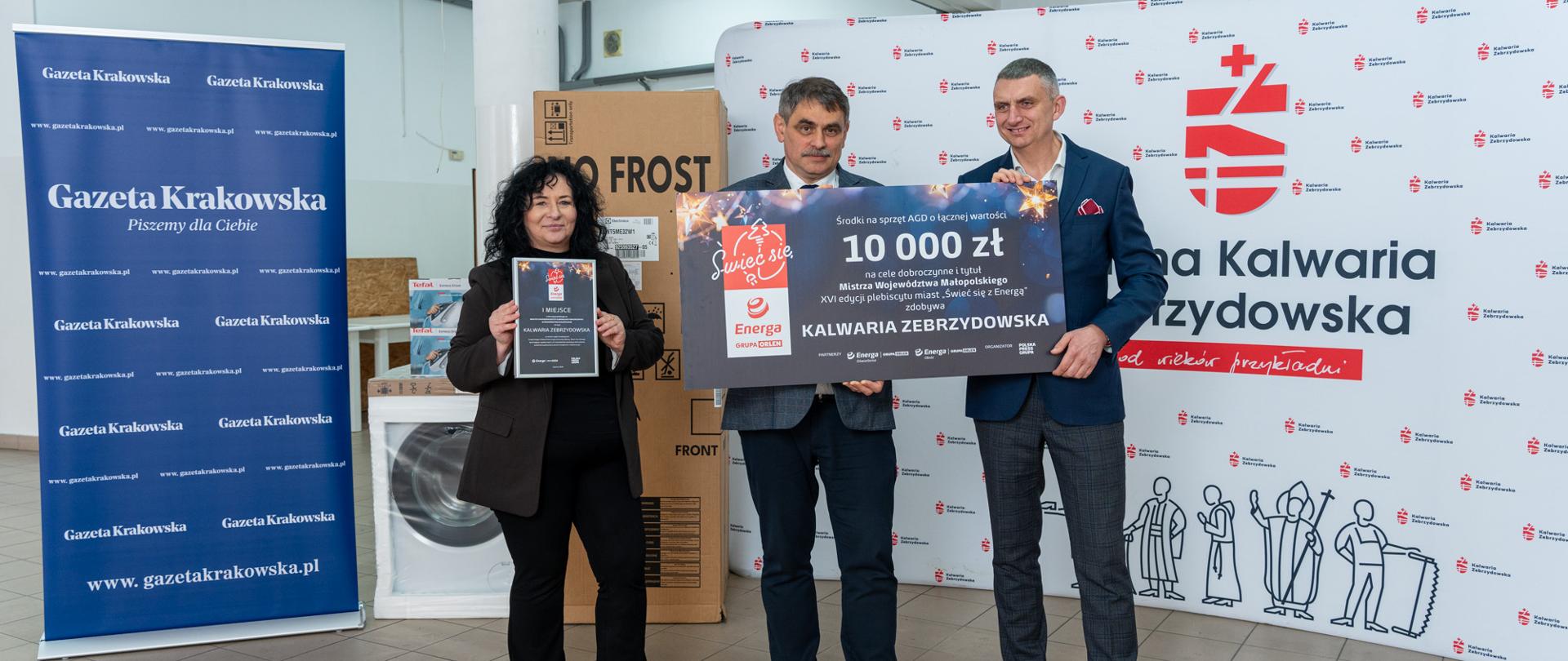 Trzy osoby stoją w pomieszczeniu urzędu na tle ścianki promocyjnej Gminy Kalwaria Zebrzydowska oraz baneru „Gazety Krakowskiej”. Dwie osoby trzymają duży symboliczny czek o wartości 10 000 zł, a trzecia prezentuje statuetkę za zwycięstwo w konkursie „Świeć się z Energą”. Obok widoczne są zapakowane sprzęty AGD, m.in. pralka i kartony z wyposażeniem przeznaczonym dla potrzebujących.