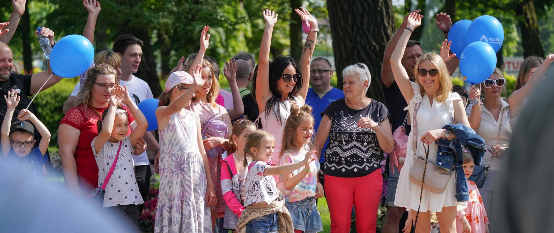 ucieszeni dorośli i dzieci podczas Dnia Dziecka stoją w grupie i podnoszą ręce. Widać balony i psa