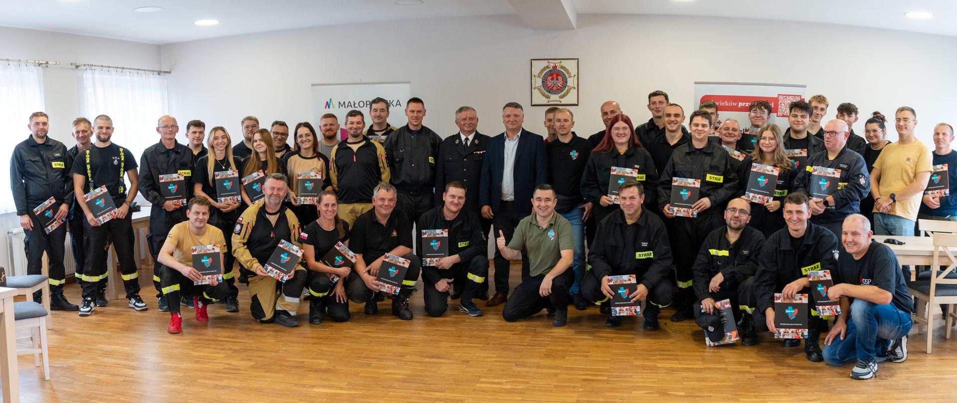 Zdjęcie grupowe uczestników szkolenia z kwalifikowanej pierwszej pomocy w Kalwarii Zebrzydowskiej. W sali pozują strażacy OSP, ratownicy, organizatorzy oraz przedstawiciele samorządu. Część uczestników trzyma materiały szkoleniowe i certyfikaty. W tle widoczne są roll-upy z logo Małopolski oraz znak straży pożarnej.
