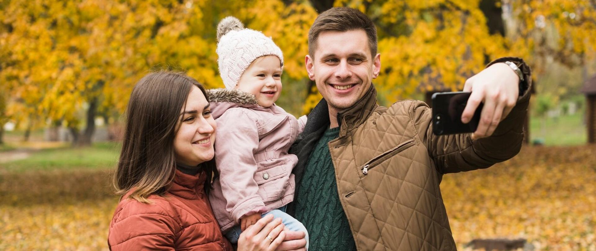 Rodzice podczas jesiennego spaceru po parku robią sobie selfie z dzieckiem na ręce, wszyscy się uśmiechają