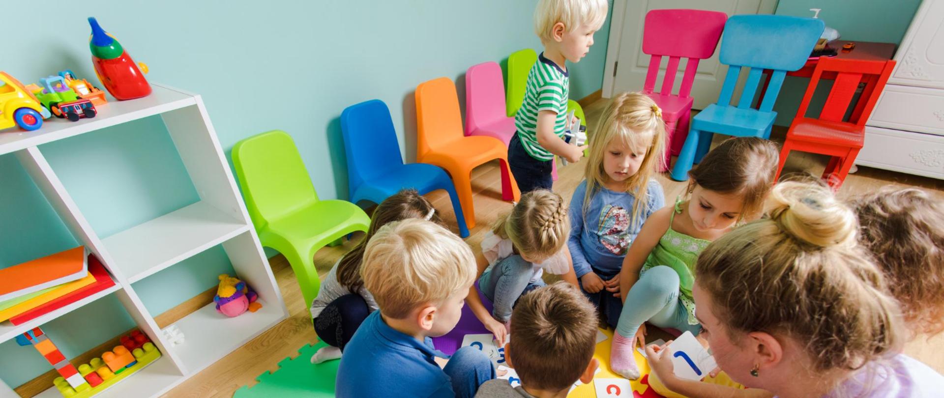 Grupa dzieci wraz z opiekunką siedzi na kolorowej macie edukacyjnej w sali przedszkolnej. Uczestnicy zajęć wspólnie układają karty z literami i uczą się poprzez zabawę. W pomieszczeniu znajdują się kolorowe krzesełka, regał z zabawkami oraz pomoce dydaktyczne dostosowane do wieku dzieci.