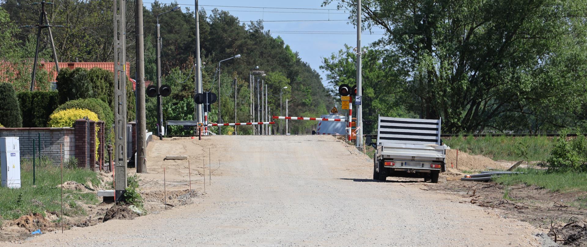 Przejazd kolejowy na drodze z kruszywa. Na przejeździe są opuszczone szlabany. Po prawej stronie stoi samochód dostawczy