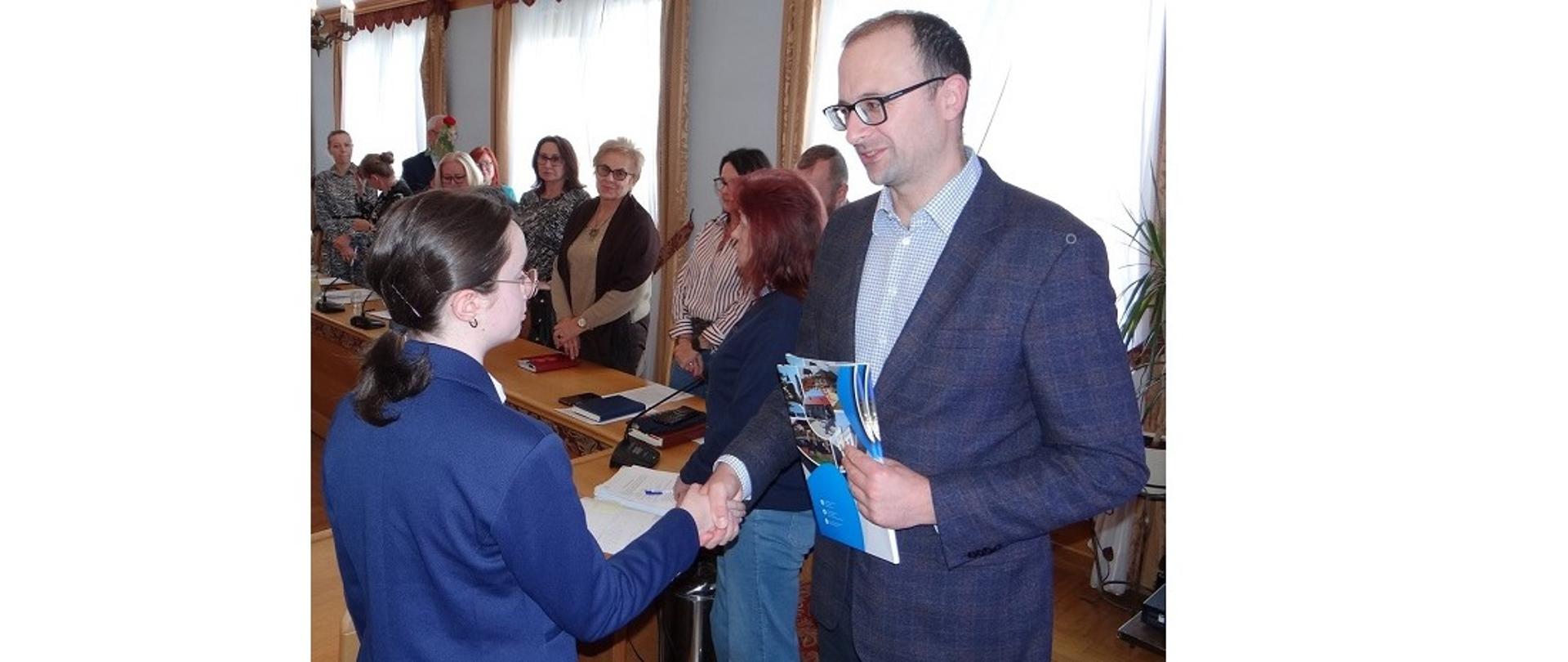 Wójt Grzegorz Sikorski gratuluje Julii Samol zdobycia stypendium. Młoda dziewczyna odbiera gratulacje przypieczętowane przez wójta uściskiem dłoni w obecności osób obecnych w sali 