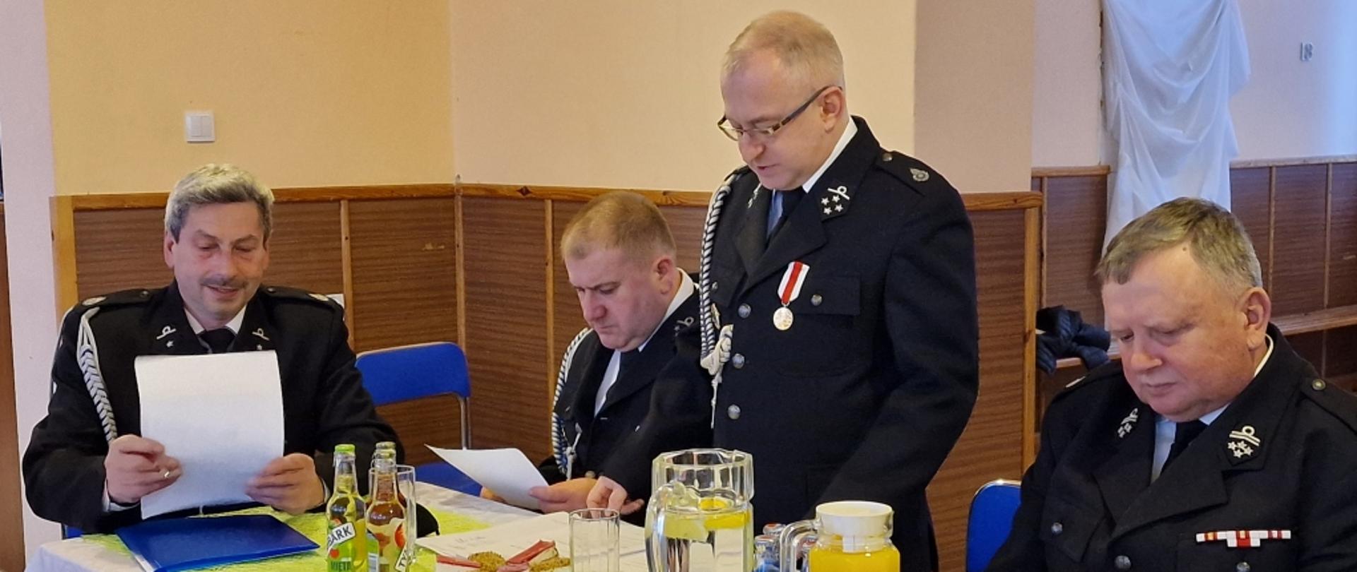 Stół prezydialny zebrania OSP Zarzyce Wielkie, na pierwszym planie stoi radny Piotr Kumor, obok siedzą strażacy ochotnicy